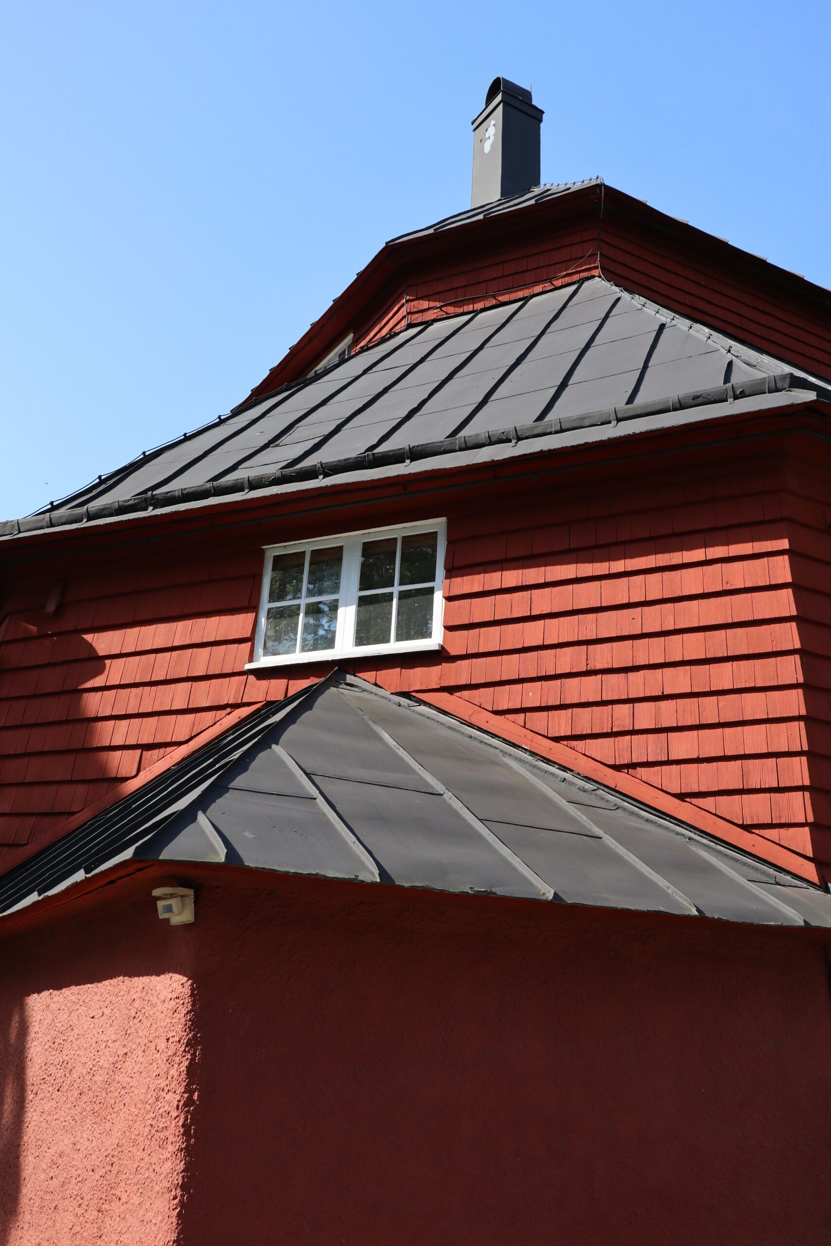 Close-up of a red building with a black roof and chimney under a clear blue sky, featuring a window.