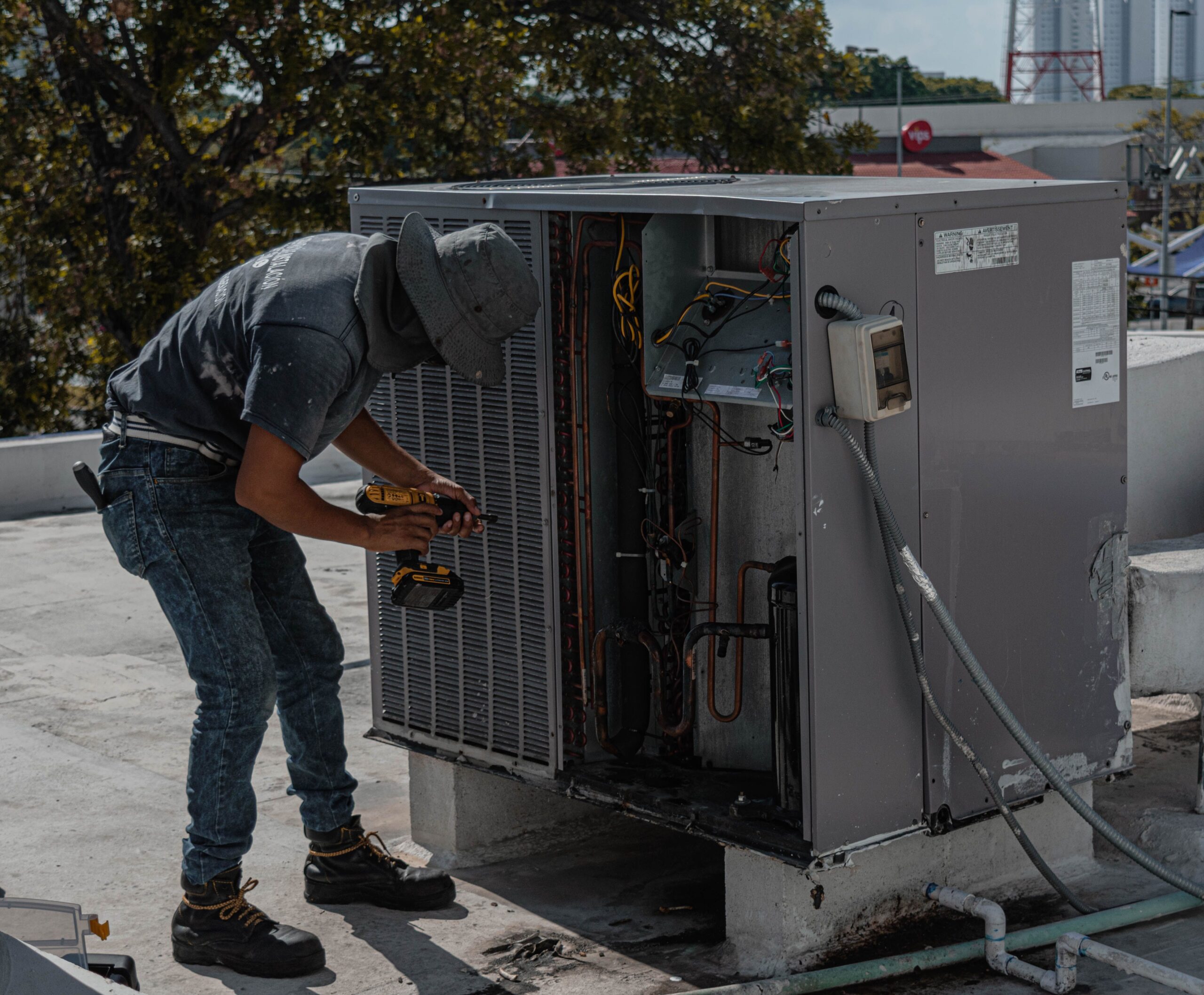A worker in a bucket hat repairs an outdoor air conditioning unit on a rooftop.